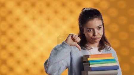 Portrait of woman with pouting expression holding pile of books, showing disproval of reading hobby. Sulky lady with stack of novels doing thumbs down hand gesturing, studio background, camera A