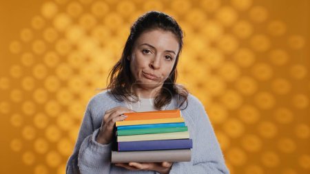 Portrait of woman with pouting expression holding pile of books, showing disproval of reading hobby. Sulky lady with stack of novels doing thumbs down hand gesturing, studio background, camera B