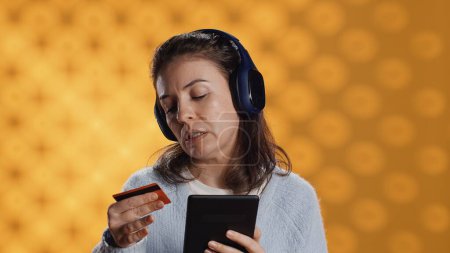 Photo for Woman buying electronic books for ereader using credit card, isolated over studio background. Bookworm listening music in headphones, purchasing ebooks, typing on tablet screen, camera A - Royalty Free Image