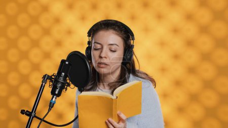 Narrator wearing headset reading aloud from book into mic against yellow background. Upbeat professional voice actor recording audiobook, creating engaging media content for listeners, camera A