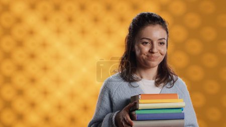Portrait of smiling woman holding stack of books, doing salutation hand gesture, feeling optimistic. Jolly person with pile of novels raising arm to greet someone, studio background, camera A