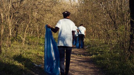 Photo for African american girl collecting rubbish in a trash bag using tongs, recycling plastic waste and picking up garbage in a forest. Young woman activist clearing the woods from junk. Camera B. - Royalty Free Image