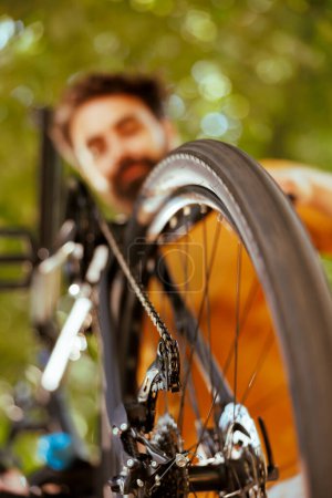 Detailed shot of bicycle wheel rubber chain being examined and maintained by young healthy caucasian man outdoor. Image showing close-up view of bike tire and pulley wheels in yard.