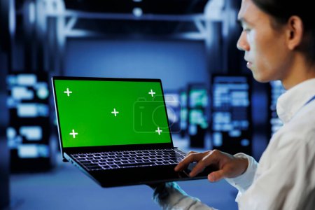 Photo for IT consultant with green screen laptop between server hub clusters providing processing resources for businesses worldwide. Worker uses mockup device to fix data center mainframes - Royalty Free Image