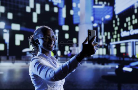 BIPOC-Frau, die nachts durch die Stadt läuft und ein Selfie mit dem Handy macht. Bürger fotografiert mit Smartphone bei Spaziergängen durch leere, von Lampen erleuchtete Straßen