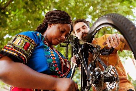 Photo for Sporty and eager black woman in yard repairing bike derailleur for summer recreational cycling. Healthy committed african american female cyclist assisted by caucasian man in repairing of bicycle - Royalty Free Image