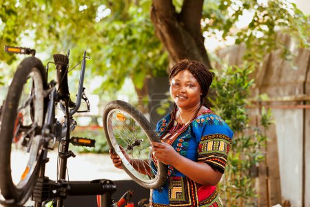 Photo for Portrait shot of energetic female cyclist holding dismantled bicycle tire for repair and maintenance in home yard. Active sporty african american woman handling bike wheel for reattachment. - Royalty Free Image
