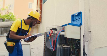 Photo for African american electrician doing leak checks and other necessary fixes to prevent major breakdowns. Skillful serviceman verifying air conditioner, writing findings on clipboard - Royalty Free Image
