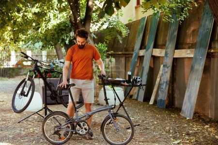 Kaukasischer männlicher Radfahrer, der in seinem Hof Hausarbeiten erledigt, umgeben von einer Sammlung professioneller Radsportausrüstung. Sportlicher Mann sorgt nach Reparatur für Sicherheit des Fahrrads.