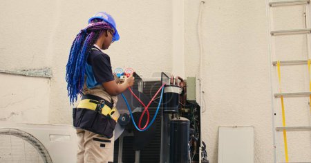 Skilled african american expert looking at freon levels in air conditioner while using manifold indicators to measure the pressure in HVAC system, ensuring efficient cooling performance
