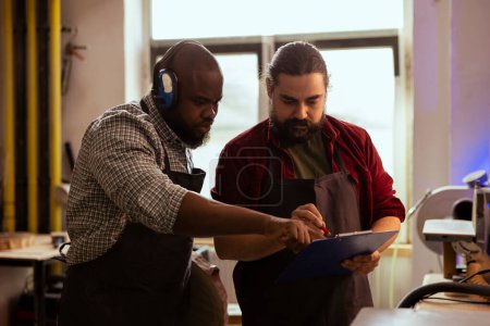 Photo for Carpenter and coworker looking over technical drawings on notepad to make creative wood art pieces. Artisan and african american man looking at blueprints to execute woodworking projects - Royalty Free Image
