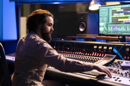 Portrait of music producer working in control room with sliders and switchers, adjusting volume level on audio recordings. Audio engineer works with mixing console in professional studio.