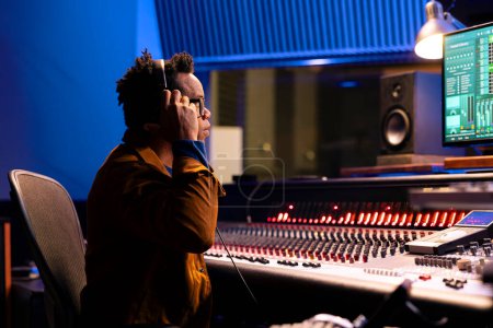 Photo for African american tracking engineer processing and editing music in studio control room, operating mixing console with moving sliders and colored meters. Audio technician deals with stereo gear. - Royalty Free Image