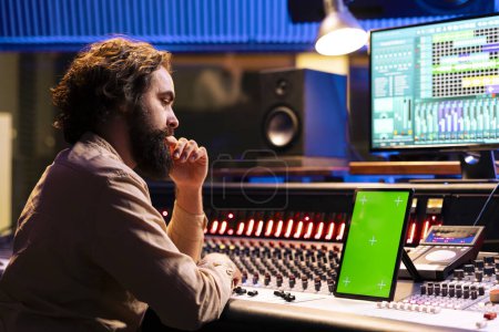 Skilled sound engineer looks at mockup display on tablet before mixing and mastering tracks on electronic stereo console. Technician uses digital audio software to record and edit his songs.