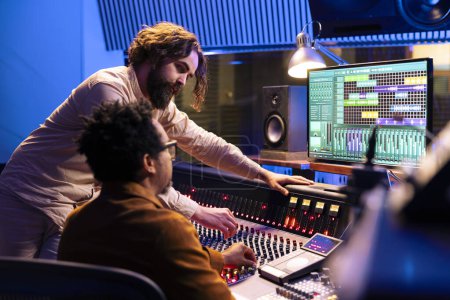 Skilled artist working with audio technician to edit recorded songs in control room, pressing sliders and levers on panel board. Team of singer and sound designer creating records in studio.