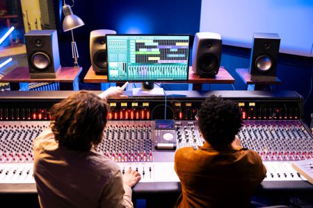 Audio engineer and artist collaborating on new songs for pop album, composing and editing tunes with mixing console and digital software. Musician working with technician on records in studio.