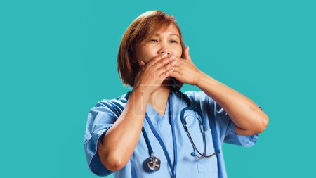 Diplom-Krankenschwester bedeckt Mund mit Händen und hält sich mit unhöflichen Gedanken zurück. Krankenhausmitarbeiterin schweigt, schweigt, schweigt, isoliert über Studiohintergrund, Nahaufnahme