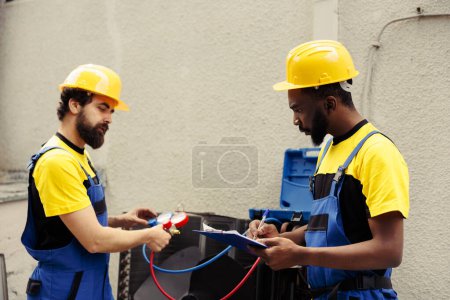 Photo for Trained engineer using set of manometers to read pressure of liquids and gases in condenser unit while expert repairman writes air conditioner faulty components report list on clipboard - Royalty Free Image