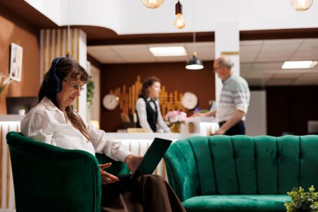 Anciana jubilada usando auriculares y sentada en un sofá sosteniendo su computadora personal. Cliente senior que tiene auriculares inalámbricos espera en el vestíbulo del hotel y utiliza el ordenador portátil para navegar por Internet.