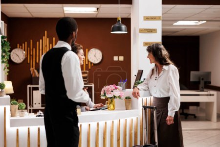 Mujer jubilada en recepción haciendo el check-in en el exclusivo vestíbulo del hotel. Mientras botones ayuda con la maleta en la sala de estar, conserje ayuda a anciana turista con reserva de habitaciones.