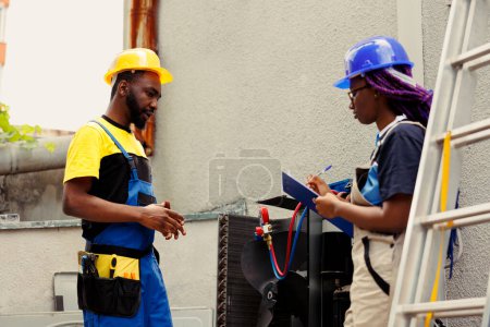 Photo for Seasoned mechanics repairing heating and ventilation of air conditioning system. Adept repairmen commissioned for external hvac system yearly maintenance, writing report on clipboard - Royalty Free Image