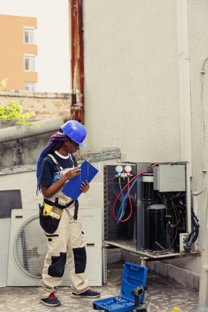 Photo for Efficient technician commissioned to optimize HVAC system performance, ensuring it operates at maximum efficiency. Seasoned mechanic doing air conditioner checkup, imputing data on clipboard - Royalty Free Image