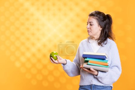 Téléchargez les photos : Femme avec pile de livres dans les mains profitant de pomme fraîche, vivant en bonne santé, isolé sur fond de studio. Ver de bibliothèque contenant une pile de romans et de fruits verts, dégustant une collation bio - en image libre de droit