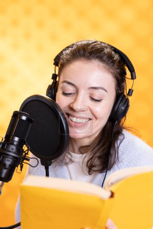 Happy narrator wearing headphones reading aloud from book into mic against yellow background. Joyous lady recording audiobook, creating engaging media content for listeners