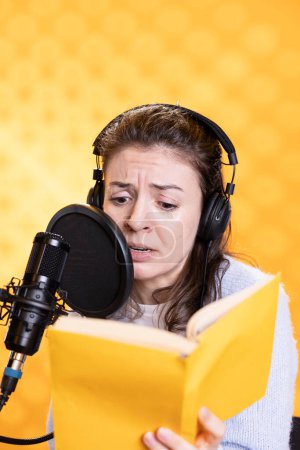 Frau mit Stirnrunzeln im Gesicht und Kopfhörern, die vor dem Hintergrund aus einem Buch in ein Mikrofon vorlesen. Sprachakteur nimmt Hörbücher auf, schafft fesselnde Inhalte für die Zuhörer, düstert für dramatische Effekte