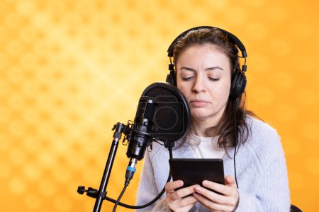 Narrator wearing headphones reading aloud from ebook on ereader into professional microphone against backdrop. Voice actor recording audiobook, reading text from tablet screen, doing audio story