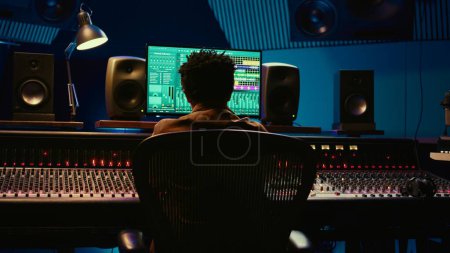 Photo for African american sound designer mixing and mastering tracks in professional studio, using audio console with moving faders and colored meters. Music producer working on control room. Camera A. - Royalty Free Image