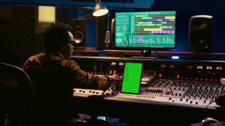 Photo for African american audio technician mixing and editing tracks with greenscreen on tablet, working in professional post production studio. Producer creating new tunes for his album. Camera B. - Royalty Free Image