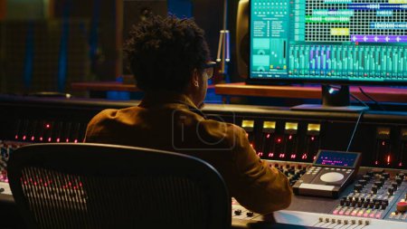 Audio expert counts down for artist singing in vocal booth, recording tracks to edit them after in post production. Producer operates mixing console and buttons or sliders, control room. Camera A.