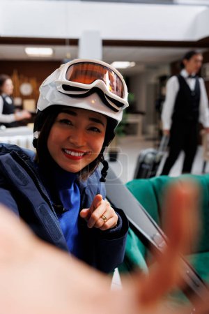 Foto de Mujer asiática joven en la exclusiva estación de esquí sosteniendo el teléfono celular para foto selfie. Mujer entusiasta viajera con chaqueta de invierno gafas de esquí y casco listo para actividades invernales. - Imagen libre de derechos