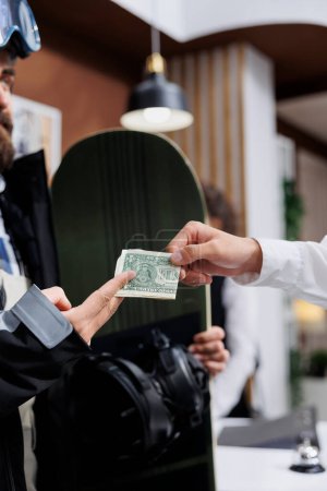 Photo for Close-up of male traveler with snowboard giving tip money to employee at counter of luxury winter resort. Detailed shot showing man wearing snowboarding gear tipping staff with cash in hotel lobby. - Royalty Free Image