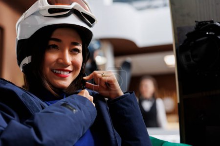 Téléchargez les photos : Femme asiatique sécurisant bande de casque d'hiver tout en étant assis dans le foyer de l'hôtel et arborant une veste d'hiver. Femme apporte des modifications de sécurité au couvre-chef de neige pour le snowboard sur les pistes de ski. - en image libre de droit