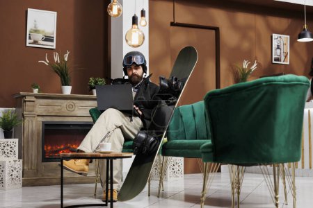 Jeune voyageur organisant des activités pour les vacances d'hiver à la station de ski de montagne sur ordinateur portable dans le salon. Homme caucasien avec équipement de snowboard naviguant sur Internet sur ordinateur personnel.