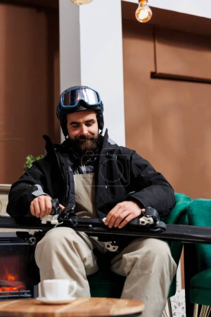 Touriste homme portant des vêtements d'hiver et casque regarde son équipement de ski en attendant la réservation dans le foyer de l'hôtel. Jeune homme assis sur le canapé saisit ses skis de ski, excité par l'aventure enneigée.