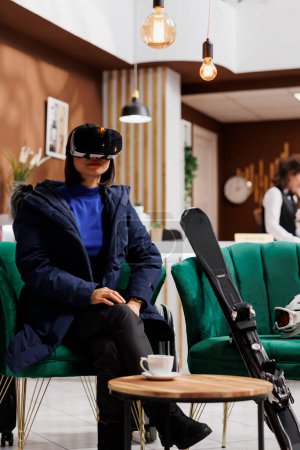 In hotel reception, young female traveler adorned in snow gear including winter jacket, attentively using vr glasses. Asian woman having vr headset sits in lounge area of ski mountain resort.