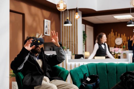 Male tourist wearing winter jacket is using a vr headset in lounge area of ski mountain resort. While waiting in hotel lobby man with beard utilizes vr goggles for research on wintersports activity.