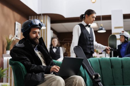 Ein kaukasischer männlicher Reisender sitzt in der Hotellobby und surft auf einem Laptop. Junger Mann in Winterjacke surft am PC im Netz und bucht Winterurlaub.