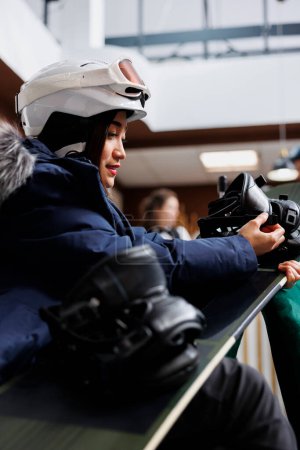 Viajero femenino listo para el invierno que se ajusta y garantiza la seguridad de las botas de snowboard en el vestíbulo del hotel. Mujer joven manteniendo equipo de snowboard, anticipando aventura invernal.