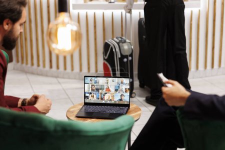 Entrepreneurs use videoconference call in hotel lobby, attending online business meeting before doing check in at reception front desk. Businessmen meet with team on video telework.