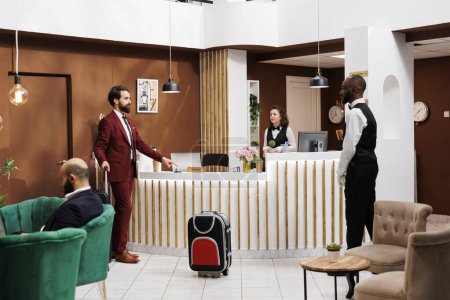 White collar worker welcomed by hotel staff at reception, carrying luggage for important business trip. Guest arriving at resort talking to receptionist and bellboy, asking about services.