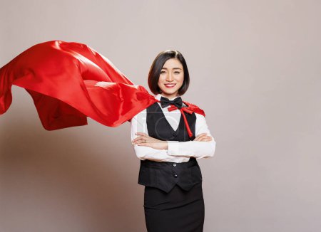 Photo for Smiling receptionist standing with folded hands wearing fluttering superwoman cloak, showing confidence and power portrait. Young asian waitress in hero red cape looking at camera - Royalty Free Image
