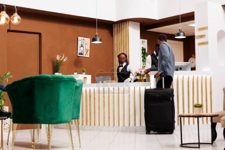 Hotel check in process. African American man traveler with suitcase standing at reception desk talking with receptionist registering his arrival, tourist waiting for key or access card to room