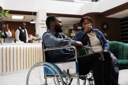 Young African American man on wheelchair sitting in hotel lobby with wife, tired unhappy woman travelling with disabled husband. Inclusive hospitality, handicap people and travel