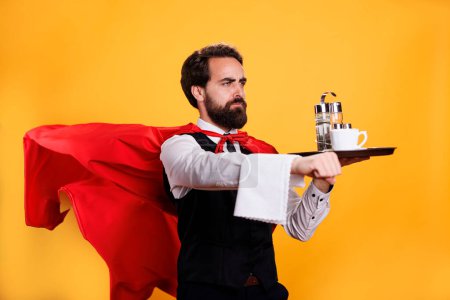 Photo for Elegant waiter with red cape carrying coffee on tray, having towel hanging over hand and posing as a superhero in studio. Young adult restaurant butler serving food and drinks. - Royalty Free Image