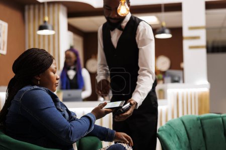 Téléchargez les photos : Femme afro-américaine invité d'hôtel assis dans le hall tenant smartphone payer pour l'ordre en utilisant la technologie nfc. Serveur tenant terminal POS pour paiement sans contact servant voyageur féminin - en image libre de droit