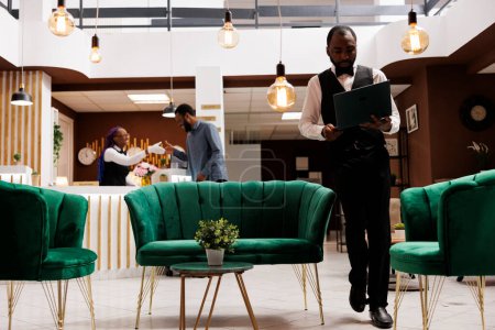 Foto de Joven hombre afroamericano empleado de hotel vistiendo uniforme de pie con el ordenador portátil en el vestíbulo, utilizando el programa de software de computadora para hacer reservas. CRM en la industria de la hospitalidad - Imagen libre de derechos